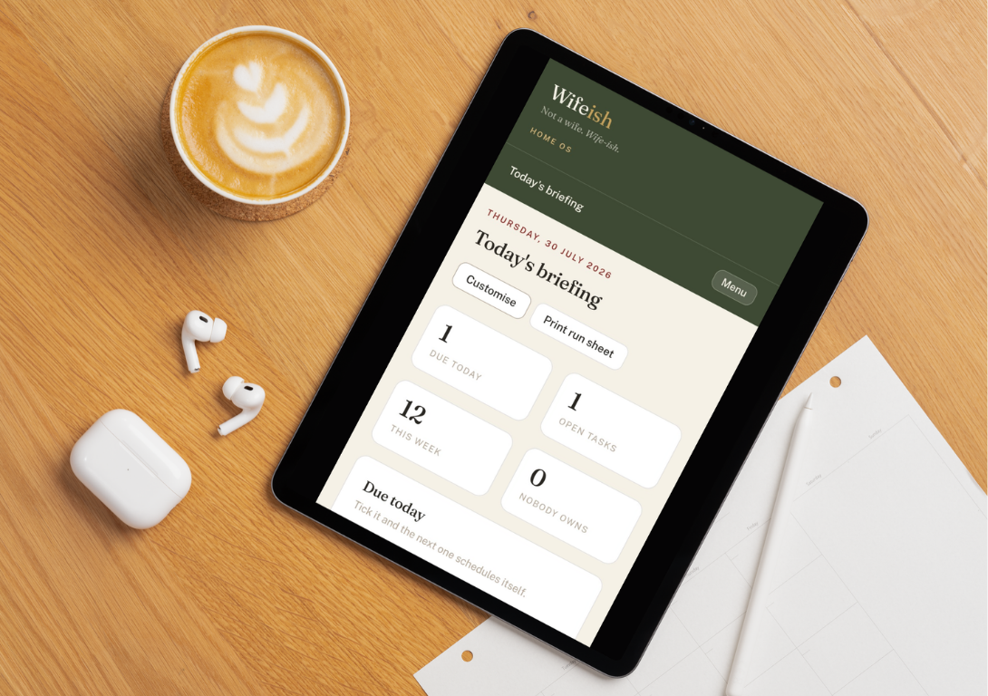 The Wifeish Home OS Today's Briefing screen shown on a tablet, resting on a wooden table beside a coffee and notepad